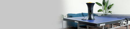 Horizontal banner image of the JOOLA Ipong trainer filled with orange table tennis balls sitting on a JOOLA table tennis table.